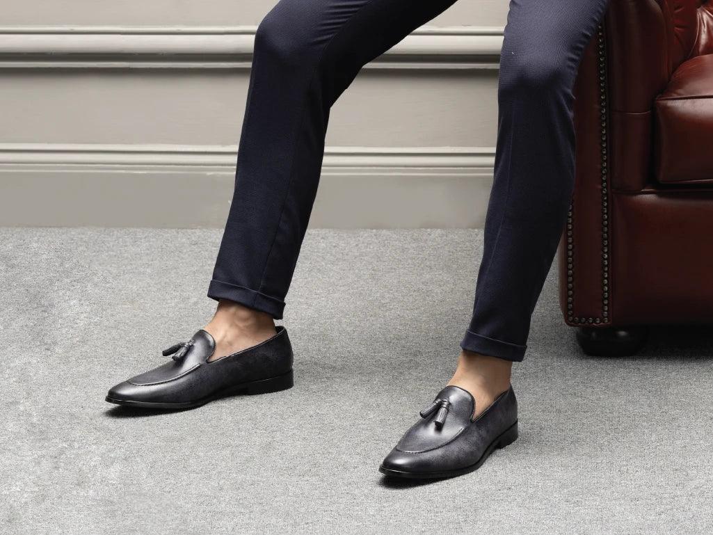 Men's black tassel loafers on grey carpet with navy pants, modern formal footwear