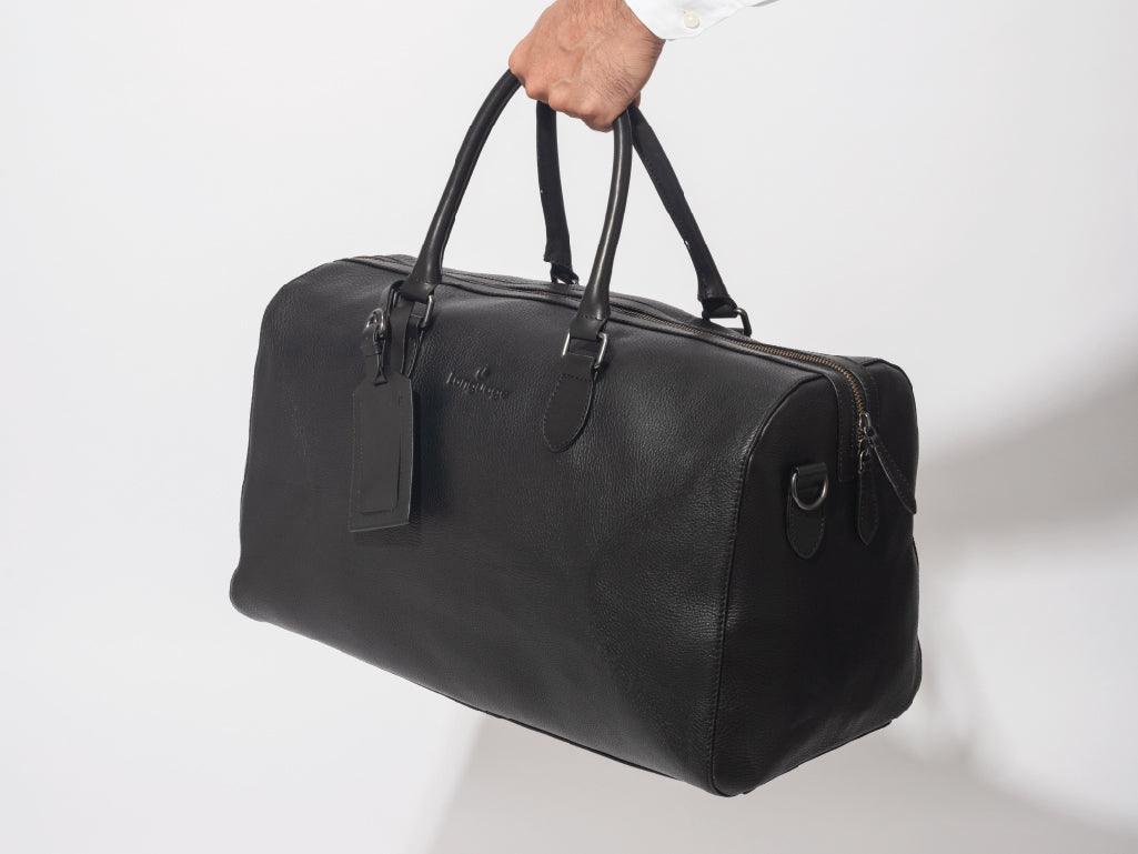 Hand holding a black leather duffel bag with top handles against a white background