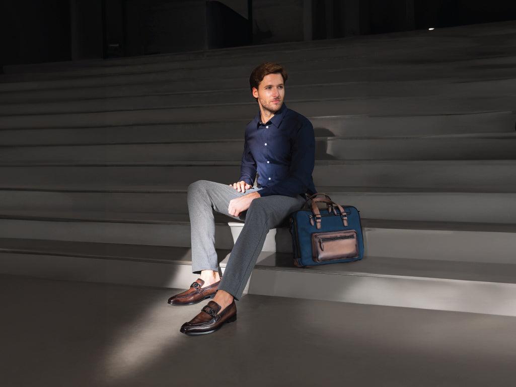 Man sitting on stairs wearing brown loafers, gray pants, blue shirt, with a leather briefcase