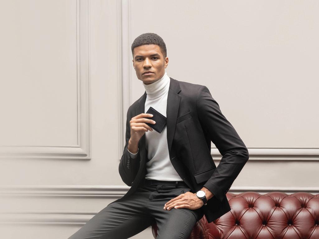 Stylish man in black suit and white turtleneck holding a wallet, sitting on a leather sofa
