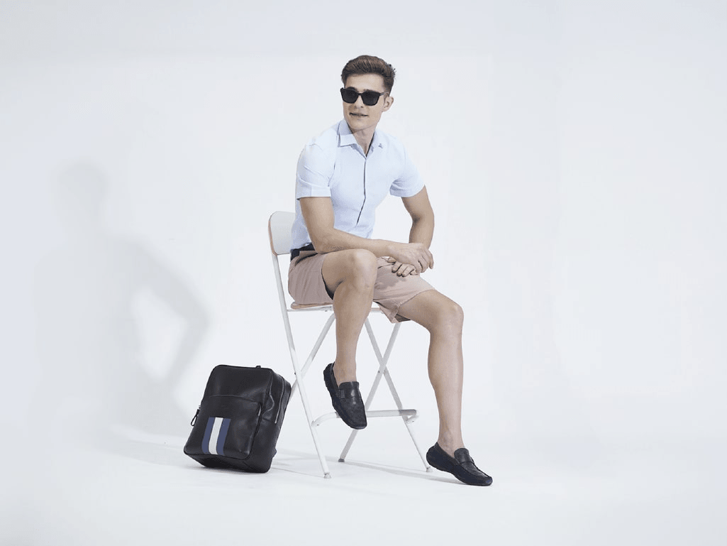 Man in sunglasses and loafers sitting on a chair with black leather backpack on floor