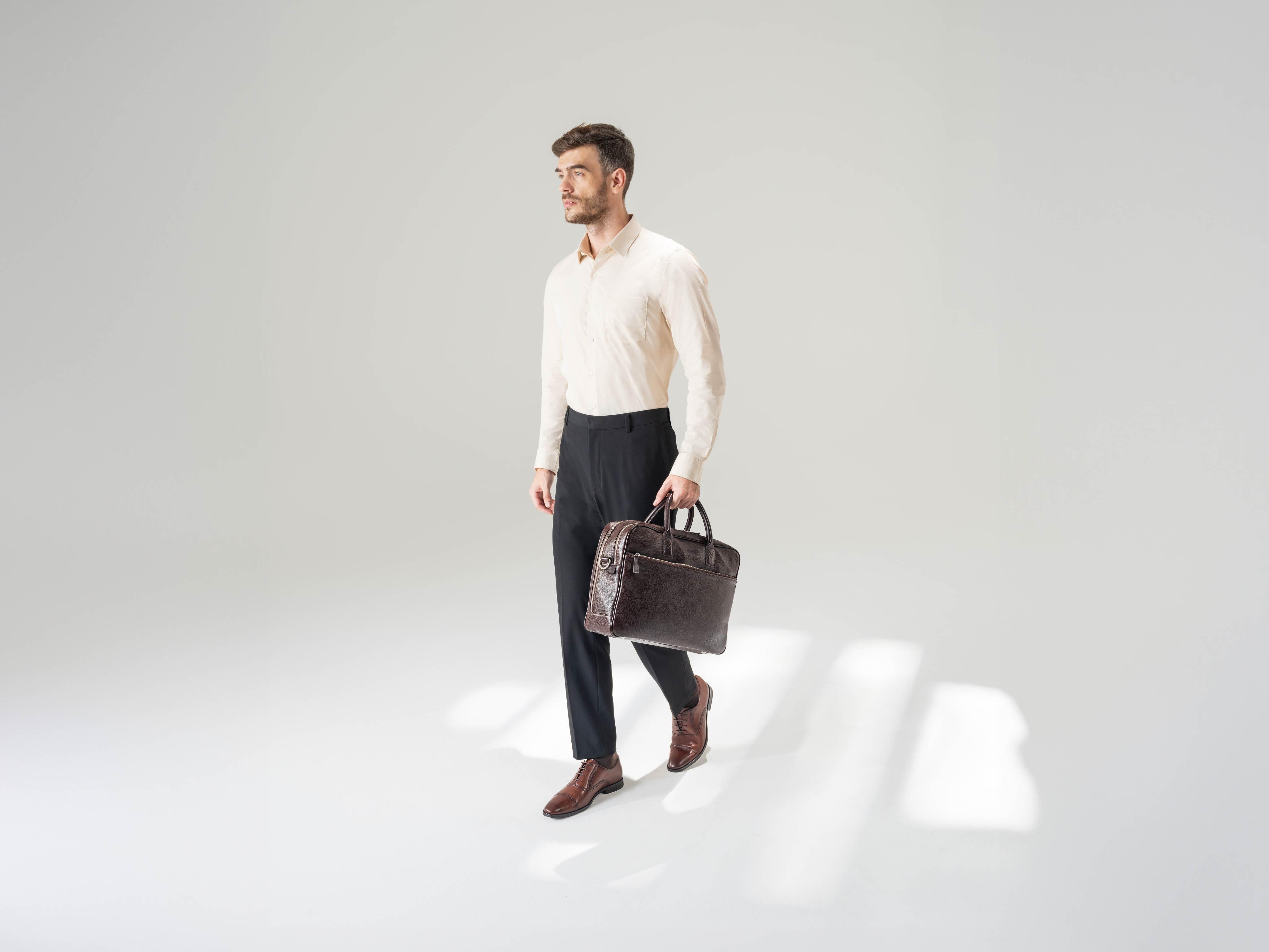 Man in formalwear carrying brown leather bag and wearing brown dress shoes on plain background