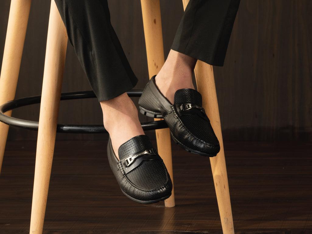 Men's black leather loafers with metal buckle detail, worn with black pants, on wooden stool