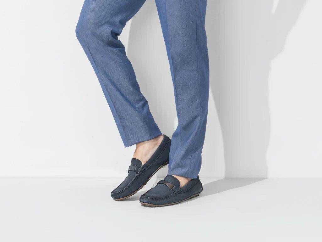 Men's navy blue loafers with stitching detail, paired with blue trousers on white background