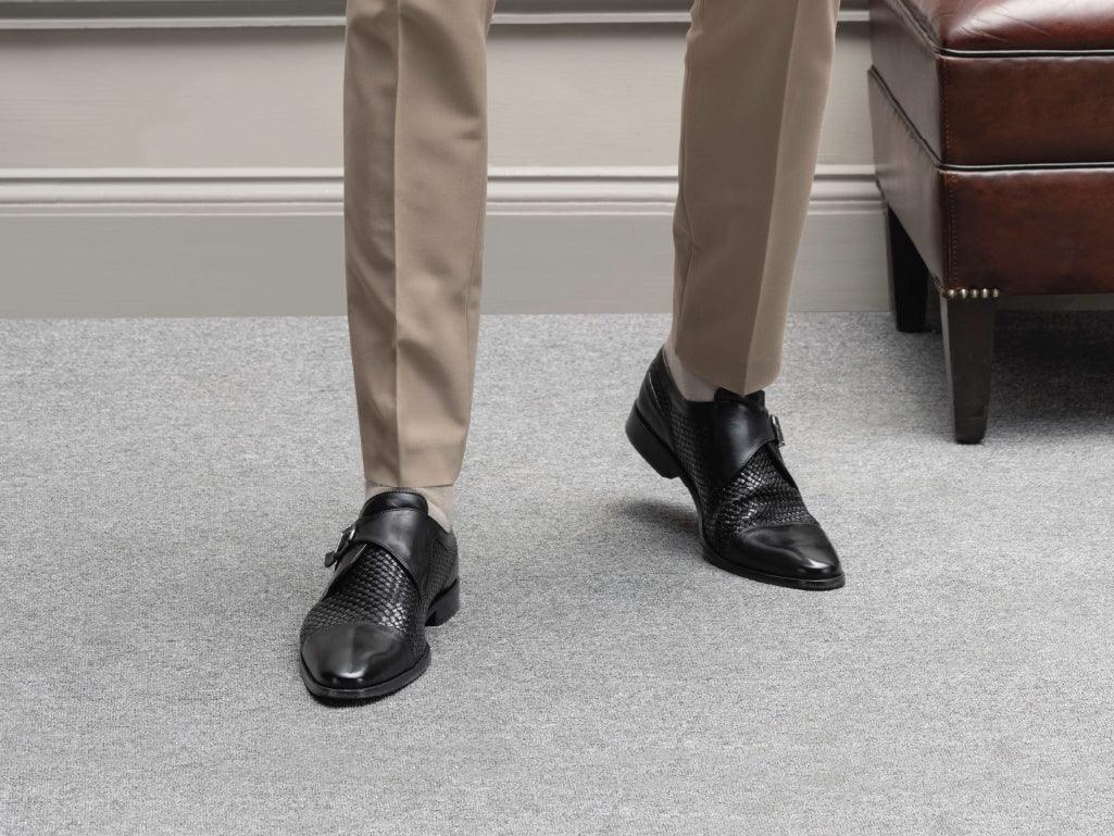 Men's black woven leather monk strap dress shoes with beige trousers on grey carpet