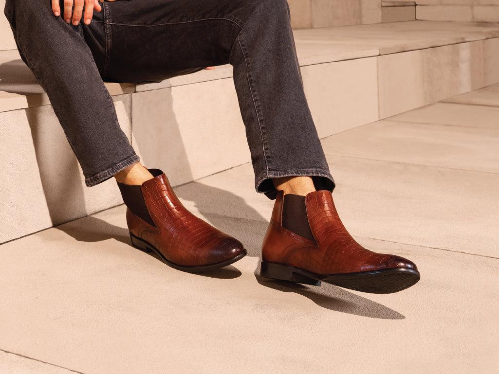 Men's brown leather Chelsea boots with crocodile pattern, worn with black jeans, outdoor setting