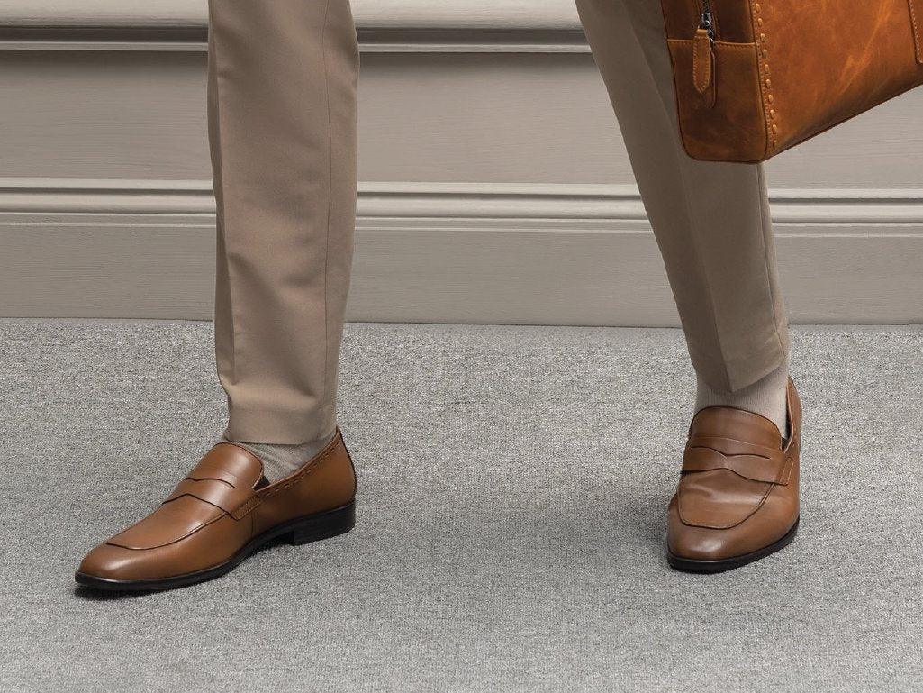 Men’s brown leather loafers with beige trousers and leather briefcase on gray carpet