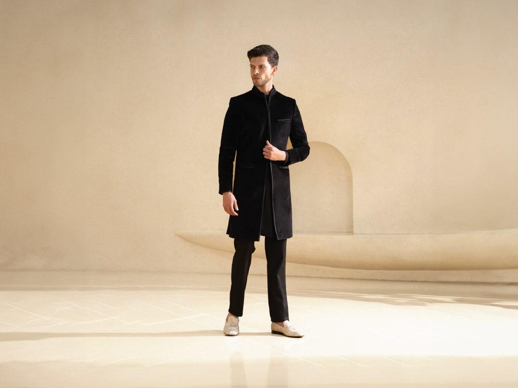Man in formal black ethnic attire and beige dress shoes standing in a minimal beige room