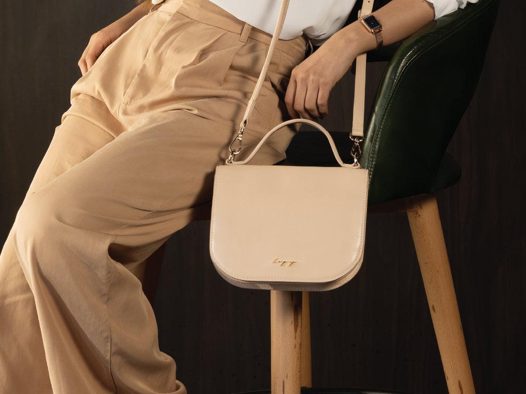 Woman seated with beige leather crossbody bag and high-waisted pants, stylish accessories