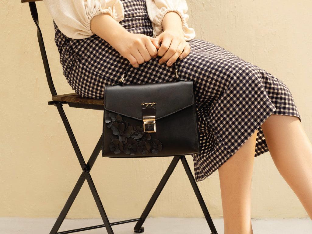 Woman sitting on chair holding black floral handbag, wearing a checkered skirt and cream blouse